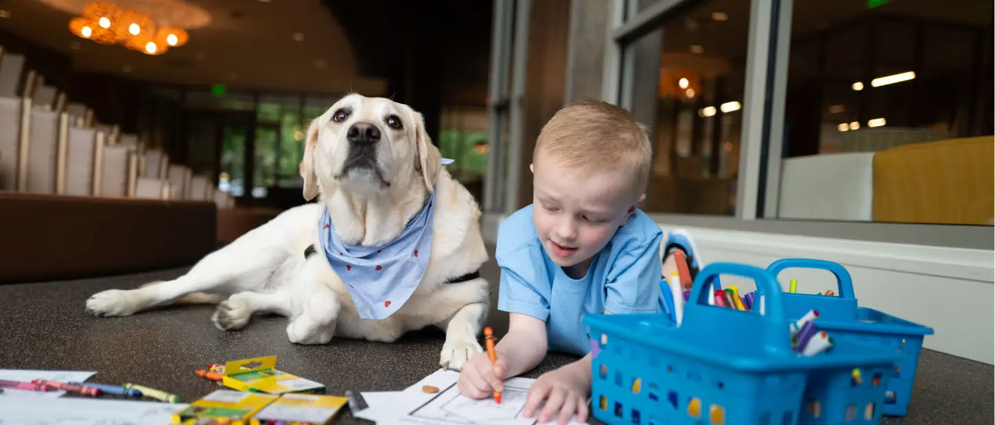 young boy sitting on ground coloring with a dog sitting closely beside