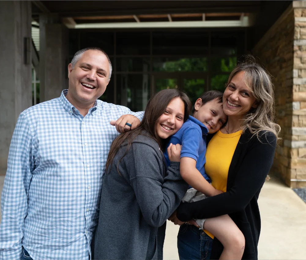a family of 4 standing in front of House smiling