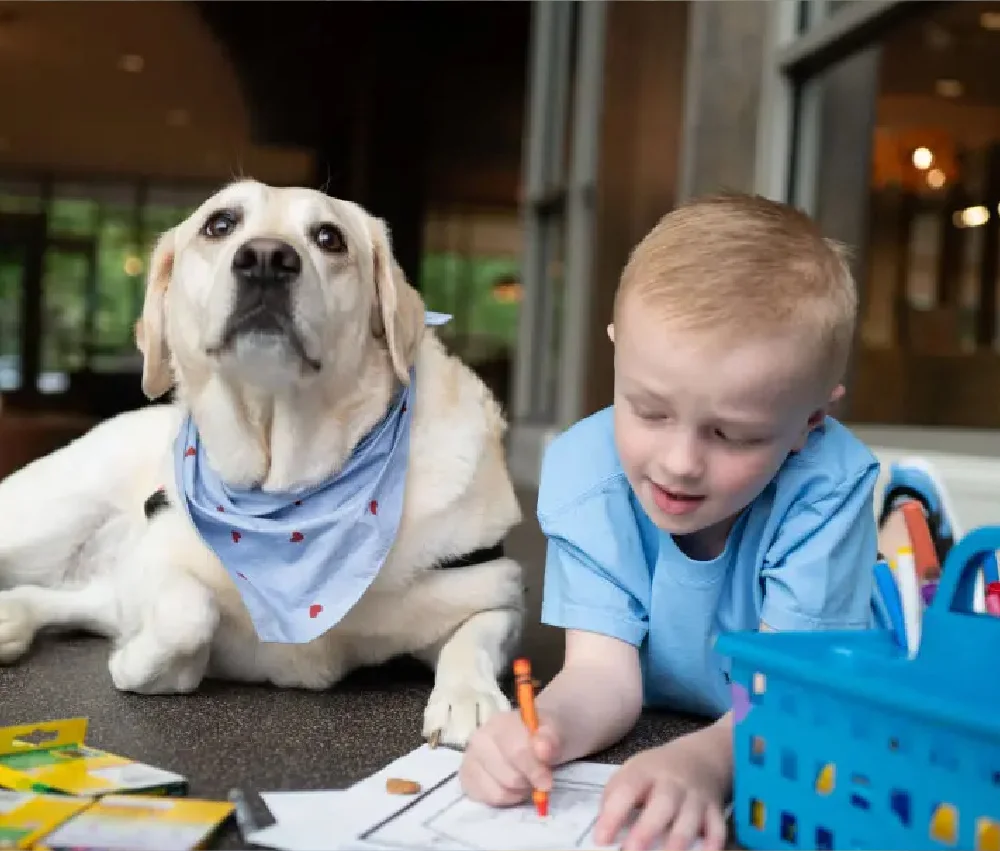 young boy sitting on ground coloring with a dog sitting closely beside